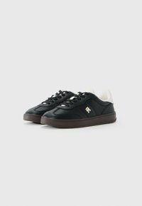 Zwarte lage sneakers met witte gevulde kraag, zwarte veters, bruine zolen en een klein wit logo aan de zijkant, tegen een witte achtergrond.