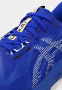 Blå idrettssko med en teksturert overdel, som har snøring, svarte detaljer og et stilisert logo-mønster i hvitt på siden.
