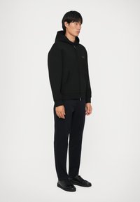 Svart hettegensere med glidelås og kengurulomme, laget av et teksturert stoff. Kombinert med marineblå bukser og svarte sneakers. Enkel design.