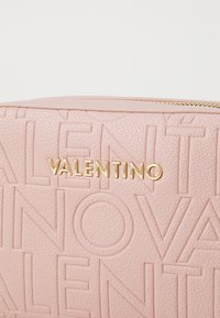 Šviesiai rožinė odinė kosmetinė, papuošta iškilmingu "VALENTINO" logotipu ir auksinės spalvos detalėmis, su užtrauktuku. Yra tekstūrinis paviršiaus raštas.