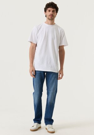 Mann steht, trägt ein weißes kurzärmeliges T-Shirt, blaue Jeans und weiße Turnschuhe vor einem schlichten hellen Hintergrund.