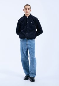 Chaqueta de pana azul marino con cierre de botones, combinada con jeans de color azul claro que presentan un patrón de texto desgastado. Unos zapatos negros completan el conjunto.