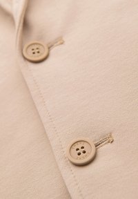 Gros plan sur un tissu beige avec deux boutons ronds beige, dont un avec le texte « OMBRE » en relief autour des trous.