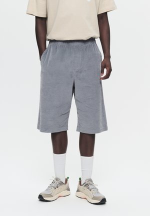 Graue Cordshorts mit Gummibund, Seitentaschen und lockerem Schnitt, kombiniert mit einem beigen T-Shirt und Sneakers.
