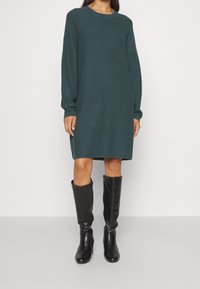 Robe pull côtelée turquoise à manches longues, coupe décontractée, arrivant au milieu des cuisses. Associée à des bottes en cuir noir jusqu'aux genoux. Texture lisse.