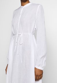 Biała sukienka z długim rękawem, zapinana na guziki, z wiązanym w pasie i miękkim, lekkim materiałem. Posiada okrągły kołnierzyk i zbierane mankiety.