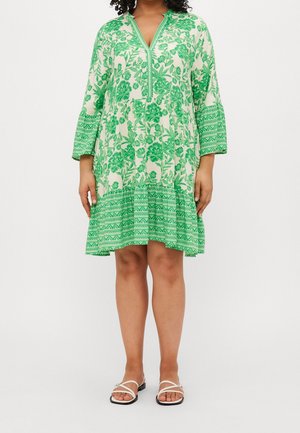 Vrouw draagt een witte jurk tot op de knie met groene bloem- en geometrische prints, gecombineerd met witte platte sandalen met bandjes.