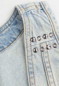 Jasnoniebieska tkanina dżinsowa ma wygięty szew na ramieniu, kontrastujące pomarańczowe szwy oraz metalowe napy wzdłuż boku do zapięcia.