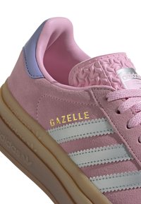 Pink suede sneaker with three white zigzag stripes, gold "GAZELLE" logo, textured pink collar, and light brown rubber sole.
