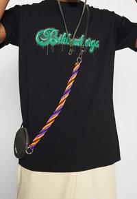 Czarna koszulka z zielonym graficznym logo, połączona z małą okrągłą czarną torbą na wielokolorowym splecionym pasku w odcieniach pomarańczowym, fioletowym i czarnym.