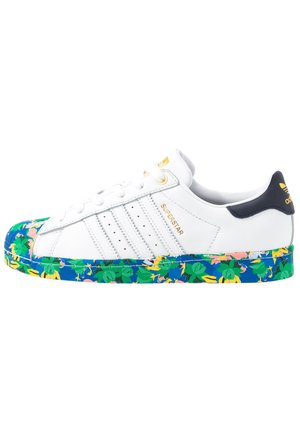 Zapatilla blanca Adidas Superstar con detalles dorados y suela azul con un estampado floral verde y amarillo.
