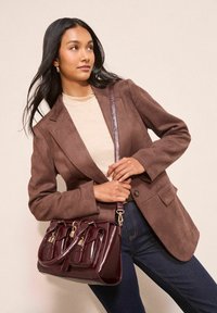 Brūns zamšs žakets, krēmkrāsas tops, tumši džinsi un spīdīgs tumši sarkans rokassomu ar vairākiem kabatām un mirdzošām detaļām.