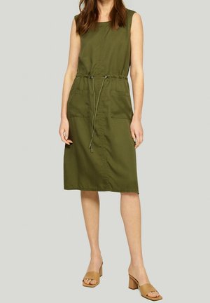 Woman wearing sleeveless olive green knee-length dress with front pockets and drawstring waist, paired with tan block-heeled sandals.