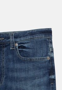 Τα σκούρα μπλε τζιν διαθέτουν τυπική μέση με θηλιές για τη ζώνη, μπροστινές τσέπες και ορατές λεπτομέρειες ραφής. Το ύφασμα έχει ελαφριά υφή.