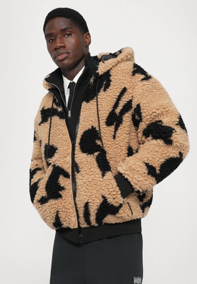 Jonge man in een beige en zwart gestreepte sherpa fleece jas met handen in zijn zakken, zwarte broek en een wit overhemd met kraag eronder.