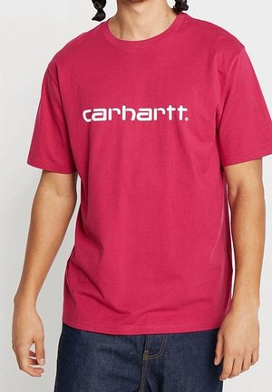 Person, die ein rotes Carhartt-T-Shirt mit kurzen Ärmeln und dunkelblaue Jeans trägt, steht vor einem schlichten hellen Hintergrund.