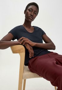 T-shirt blu navy con scollatura a barca, abbinato a pantaloni bordeaux. Il modello è seduto su una sedia di legno chiaro con seduta intrecciata.