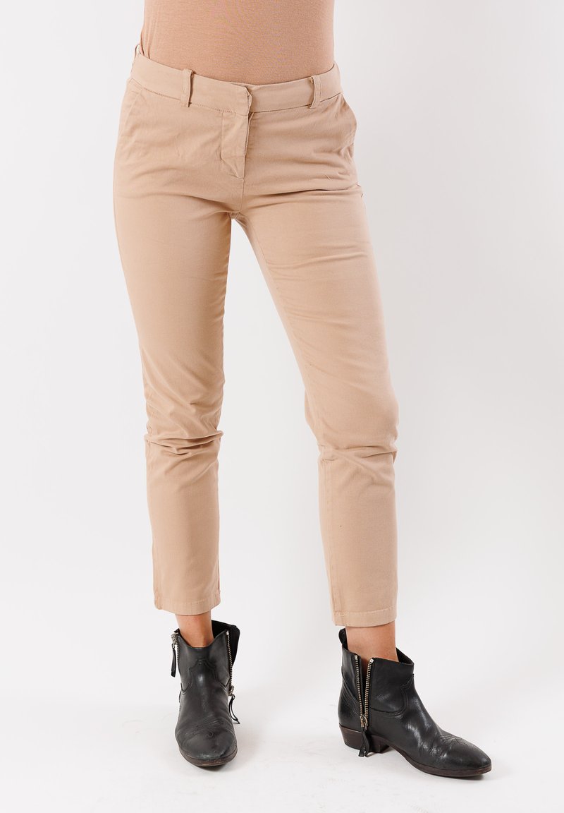 Parte inferiore indossante pantaloni beige slim-fit e stivali neri alla caviglia con cerniere laterali, in piedi su uno sfondo bianco.