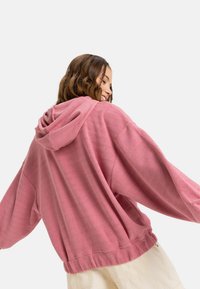 Pink hooded sweatshirt made from textured fabric, featuring a herringbone pattern, wide sleeves, and a loose fit, with a casual design.