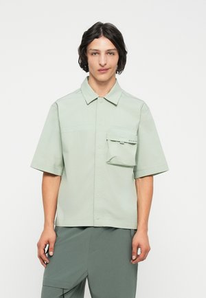 Svetlo zelena kratka majica s kratkimi rokavi, izdelana iz gladkega materiala, z gumbi spredaj in žepom na prsih z zapiranjem na pokrov.