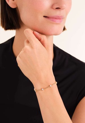 Bracciale in oro con piccole pietre preziose quadrate e trasparenti. Maglie in metallo lisce con un design delicato. Indossato su una mano che riposa sotto il mento.
