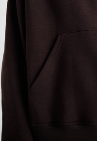 Hoodie castanho escuro com um bolso canguru, punhos e bainha canelados. Textura de tecido suave e design limpo com detalhes mínimos.