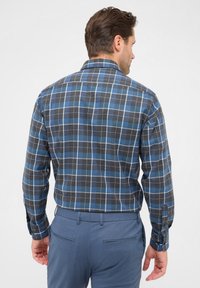 Camicia a scacchi nei colori blu, nero e bianco, con maniche lunghe. Il materiale sembra morbido, presenta un colletto posteriore con bottoni e una vestibilità rilassata.