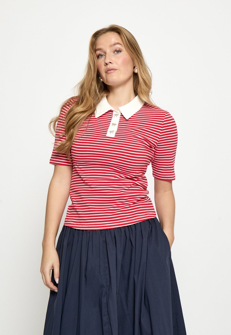 Rot-weiß gestreiftes Polo-Shirt mit kurzen Ärmeln, weißem Kragen und drei Knöpfen am Ausschnitt, kombiniert mit einem marineblauen, plissierten Rock.