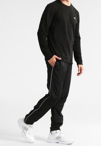Black long-sleeve t-shirt with a small logo, paired with black track pants featuring white side stripes, worn with chunky white sneakers.