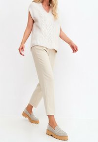 Pull crème en maille torsadée avec col en V, pantalon beige clair, et mocassins gris avec une semelle épaisse de couleur fauve. Textures lisses et coupe décontractée.