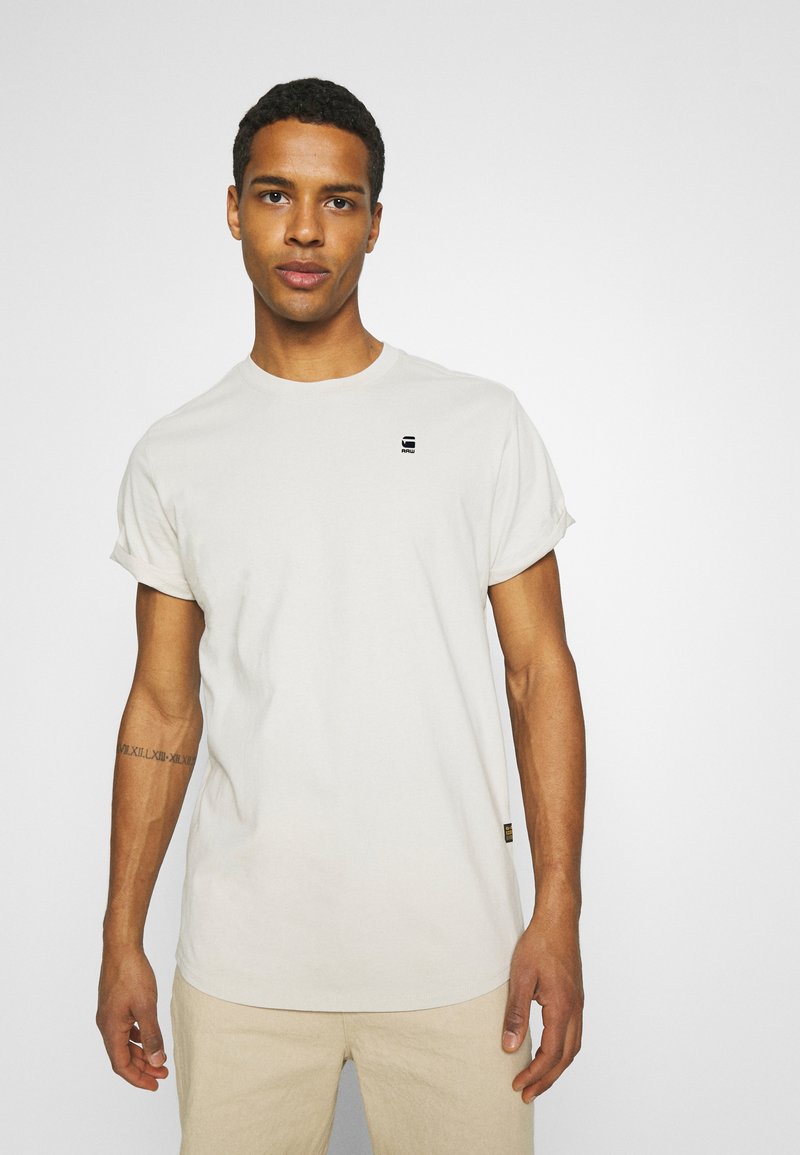 Lys beige t-skjorte laget av bomull, med rullede ermer og en liten svart logo på brystet. Enkel design med rund hals.