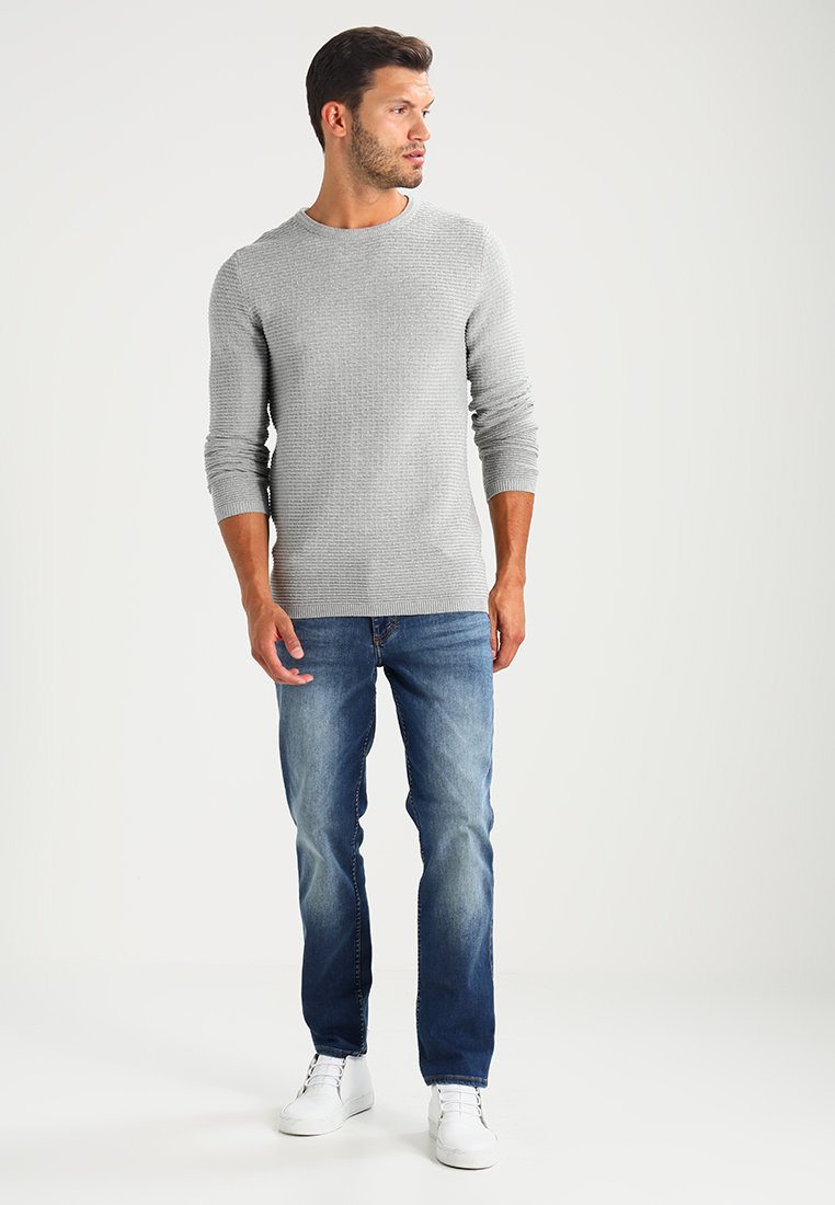 Grauer strukturierter Strickpullover mit rundem Halsauschnitt, kombiniert mit ausgebleichten blauen Jeans und weißen Sneakers, vor einem neutralen Hintergrund stehend.