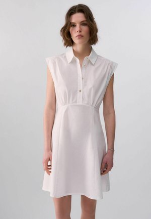 Donna con capelli castani corti che indossa un vestito bianco senza maniche con colletto e bottoni davanti, in piedi davanti a uno sfondo chiaro e semplice.