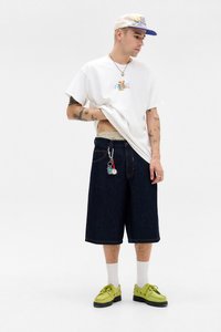 Hvid bomulds t-shirt med et farverigt grafisk design, mørkeblå denimshorts, gul-grønne sko, hvide sokker og en lys kasket med en blå skygge.