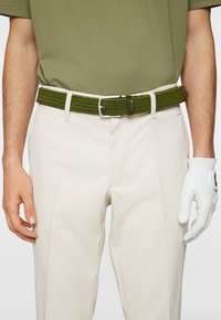 Cintura intrecciata verde con una fibbia in metallo argentato, indossata sopra pantaloni beige. Un guanto da golf bianco è visibile sulla mano destra.