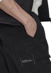 Svarta byxor med en mjuk textur, sidoficka och en liten vit tryckt etikett med text på nedre benet. Avslappnad design, funktionell stil.