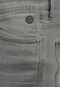 Grå denimjeans med en subtil tekstur, en svart preget knapp, og to frontlommer med kontrasterende sømdetaljer.