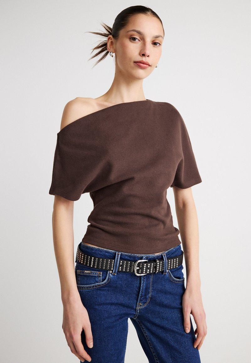 Καφέ τοπ με ακάλυπτους ώμους από πλεκτό ύφασμα, συνδυασμένο με ψηλόμεσο μπλε τζιν και μαύρη ζώνη με καρφιά. Κομψός σχεδιασμός και υφή.