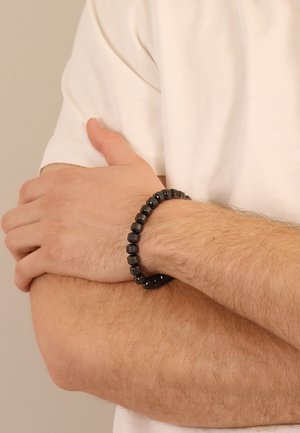 Schwarzes Perlenarmband mit glatten und texturierten Perlen, am Handgelenk getragen. Das Material scheint aus Stein oder Harz zu sein, mit verschiedenen Oberflächen.