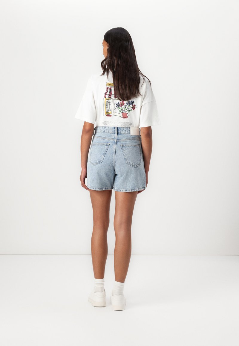 Weißes T-Shirt mit einer illustrierten Design, das ein Gebäude und Blumen auf der Rückseite zeigt, kombiniert mit hellblauen Jeansshorts.