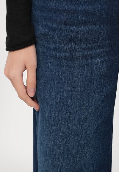 Temno modra jeansa tkanina z gladko teksturo in subtilnim bledenjem. Roka počiva ob tkanini, prikazuje podrobnosti od blizu.