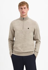 Beige gestrickter Pullover mit einem mit Reißverschluss versehenen hohen Kragen, Rippbündchen und Saum. Mit einem kleinen schwarzen Sechseck-Logo auf der Brust.