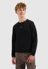 Tiener met tousled blond haar, gekleed in een zwart sweatshirt met de tekst "BALLIN", bruine broek, die iets naar de zijkant kijkt op een grijze achtergrond.