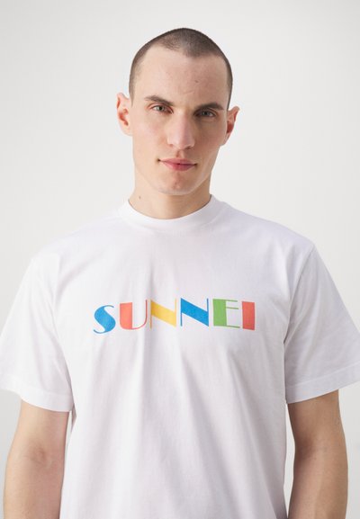 White cotton t-shirt featuring the word "SUNNEI" in bold, multicolored letters (blue, yellow, red, green) with a crew neckline and short sleeves.
