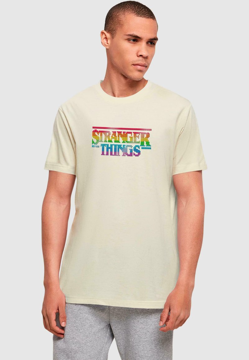 Béžové bavlněné tričko s vícebarevným grafikou "STRANGER THINGS" v tučném, pixelovém písmu přes hruď. Krátké rukávy, kulatý výstřih.