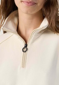 Beige ritsweater met hoge kraag, voorzien van een zwarte ritssluiting en een glad, textuurstof. Close-up van de halslijn.