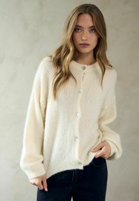 Kremai balta kardigāna augšdaļa izgatavota no pūkaina materiāla, ar apaļu izgriezumu, sudrabainiem pogām un platām piedurknēm. Valkāta pāri tumšiem džinsiem.
