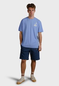 Ljusblå bomullströja med korta ärmar och en bröstgrafik, tillsammans med mörk denimshorts och beige sneakers med randiga strumpor.