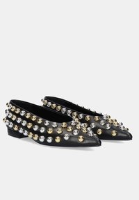 Zapatos planos de cuero negro con puntas afiladas, adornados con tachuelas doradas y plateadas en un diseño estampado, con un tacón bajo.