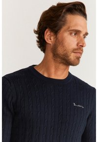 Mörkblå stickad tröja med en texturerad kabelmönster, rund halsringning och liten broderad logotyp på bröstet.